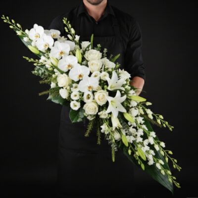 Dessus de Cercueil "Majesté Blanche" ​L'Esprit de la Création ​Une composition monumentale et d'une élégance absolue, conçue pour un hommage d'exception. Cette pièce se distingue par l'utilisation de fleurs prestigieuses qui apportent une aura de sérénité et de grandeur. Le blanc pur, symbole de lumière et de paix, est ici sublimé par des fleurs aux lignes graphiques et sculpturales, créant un dernier adieu d'une rare distinction. ​Détails de la Composition ​Cette pièce de haute floristerie est réalisée avec des variétés d'exception : ​Dimensions : Une envergure majestueuse d'environ 1m70 de long, offrant une présence imposante et solennelle sur le cercueil. ​Fleurs de Prestige : De magnifiques orchidées Phalaenopsis blanches qui apportent une touche de luxe et de délicatesse, accompagnées de Lys royaux pour leur parfum subtil et leur prestance. ​Cœur de Bouquet : Une sélection de roses blanches impeccables et de germinis blancs pour une densité et une texture parfaite. ​Lignes & Structure : Un travail sur la verticalité avec des hampes de fleurs de complément blanches et un feuillage exotique effilé qui souligne la longueur exceptionnelle de la pièce.