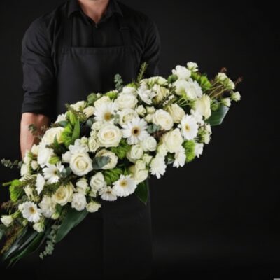 Dessus de Cercueil "Pureté Éternelle" ​L'Esprit de la Création ​Une composition majestueuse et d'une grande sobriété, conçue pour rendre un hommage empreint de sérénité et de lumière. Ce dessus de cercueil privilégie l'élégance intemporelle du blanc immaculé, symbole de paix et de respect. Sa forme allongée et son travail de texture apportent une présence réconfortante et distinguée lors de la cérémonie. ​Détails de la Composition ​Cette pièce de prestige est réalisée avec une attention particulière portée aux finitions par nos artisans : ​Dimensions : Une envergure généreuse d'environ 1m50 de long sur 35 cm de large, offrant une couverture harmonieuse et équilibrée. ​Fleurs Majeures : Une profusion de roses blanches nobles aux boutons généreux, accompagnées de germinis blancs éclatants pour la luminosité. ​Cœur de Composition : Un mélange délicat de fleurettes blanches apportant de la légèreté et du volume à l'ensemble. ​Écrin Végétal : Un travail sur le feuillage incluant de l'eucalyptus aux nuances argentées et des notes de vert profond pour souligner la pureté des fleurs.