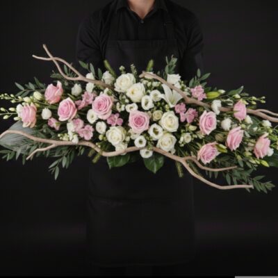 Dessus de Cercueil "Éclat de Tendresse" ​L'Esprit de la Création ​Une composition solennelle et d'une grande élégance, conçue pour rendre un hommage prestigieux et tout en douceur. Ce dessus de cercueil se distingue par sa forme allongée et aérienne, mêlant la pureté du blanc à la délicatesse des nuances rosées. Le travail des branchages naturels apporte une dimension organique et sereine à cette pièce maîtresse. ​Détails de la Composition ​Cette pièce de prestige est réalisée sur mesure par nos artisans fleuristes : ​Dimensions : Une envergure généreuse d'environ 1m50 à 1m80 de long sur 35 cm de large, idéale pour couvrir majestueusement le cercueil. ​Fleurs Majeures : Un mélange harmonieux de roses blanches nobles, de roses roses au grain lisse et de roses Bonny aux pétales délicats. ​Cœur de Composition : Des germinis et alstroemerias dans les tons blancs et roses pour assurer une densité florale parfaite. ​Structure & Style : L'ensemble est rythmé par des branchages naturels sinueux et un feuillage varié (eucalyptus, graminées) qui confèrent au bouquet sa volubilité et sa légèreté. ​Le Conseil de l'Artisan ​"Pour une telle pièce, nous sélectionnons des fleurs à différents stades d'épanouissement afin que la composition reste parfaite tout au long de la cérémonie. Nous recommandons de commander cette pièce au moins 48 heures à l'avance pour nous permettre de sélectionner les plus beaux fleurissements dans nos arrivages." ​Service & Livraison ​Cette pièce de deuil peut être commandée et coordonnée depuis nos deux magasins : ​Escapade Florale - Roncq : 10 rue Jules Cornard, 59223 Roncq. ​Escapade Florale by Sam - Wervicq-Sud : 51 rue Gabriel Péri, 59117 Wervicq-Sud. ​Livraison : Nous assurons la livraison directement sur le lieu de cérémonie ou au funérarium dans les secteurs de Roncq, Wervicq-Sud et les communes limitrophes.