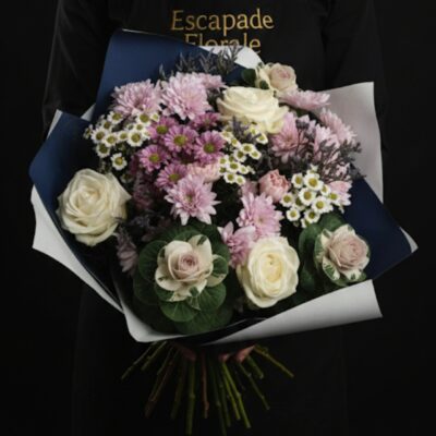 Bouquet "Douceur Champêtre" ​L'Esprit de la Création ​Une composition qui allie parfaitement la délicatesse des tons poudrés à la structure singulière des végétaux de saison. Ce bouquet rond offre un jeu de textures fascinant, mêlant la douceur des roses à l'aspect graphique du chou d'ornement. C'est une attention idéale pour célébrer une naissance, un anniversaire tout en douceur, ou pour apporter une touche de sérénité à votre foyer. ​Détails de la Composition ​Chaque bouquet est une pièce unique, assemblée avec soin dans notre atelier  : ​Pièces Maîtresses : Des choux d'ornement (Brassica) aux cœurs rosés, véritables signatures visuelles de cette composition. ​Fleurs Majeures : De magnifiques roses blanches immaculées apportant lumière et élégance. ​Cœur de Bouquet : Un mélange généreux de Bonny rose, de Santini roses et de Santini blancs pour un volume vaporeux et une harmonie de couleurs tendres. ​Finitions : Un feuillage délicat et quelques touches de fleurs de complément mauves pour souligner les contrastes.